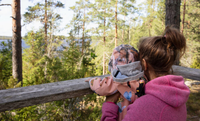 Ein Elternteil hält ein kleines Kind hoch, das auf eine sonnige Waldlandschaft blickt.