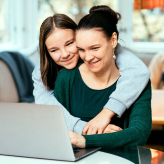 Mãe e filha sorriem sentadas em frente a um computador em sua casa.