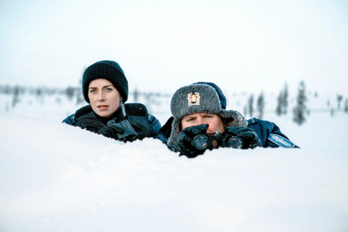 Una mujer y un hombre con gorros de invierno vigilan ocultos tras un montículo de nieve.