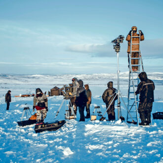 In a snowy, hilly landscape, people in thick winter clothing are setting up cameras and ladders.