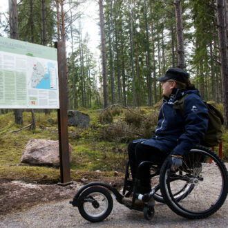 A man wearing outdoorsy clothing and a backpack sits in a three-wheeled wheelchair looking at a trailside information panel in the woods.