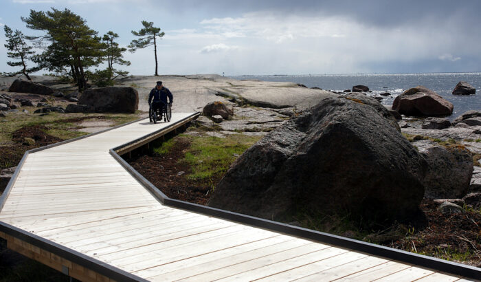 A man in a wheelchair moves along a boardwalk path that leads to a large smooth stone area on the shore.