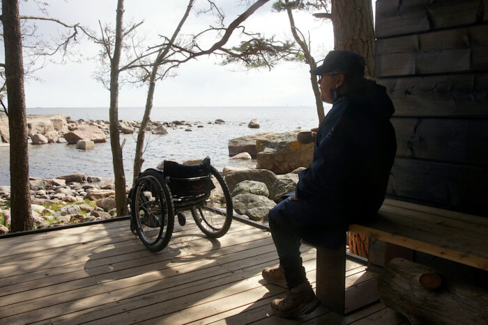 A man sits on a bench on a wooden seaside terrace, with an empty wheelchair a couple metres away from him.