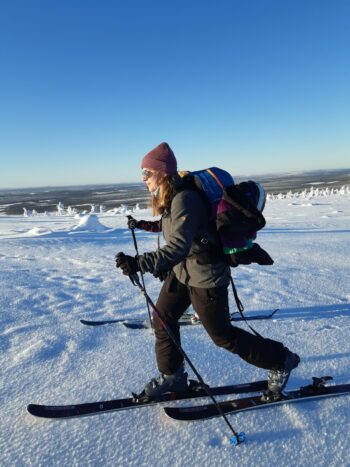 Une femme fait du ski de fond par une journée ensoleillée tandis que des sommets montagneux sont visibles à l’arrière-plan. 