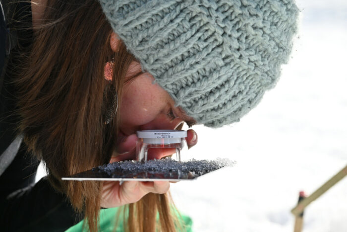 Eine Frau mit Wintermütze starrt durch eine Linse, die nahe auf ein Tablett gerichtet ist, auf Schneekristalle, die sich dort befinden.