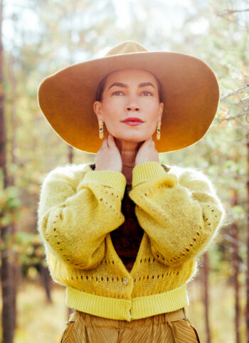 Une femme portant un chapeau à larges bords et un cardigan jaune prend la pose dans une forêt.