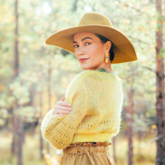 Eine Frau mit breitkrempigem Hut und gelber Strickjacke posiert in einem Wald.