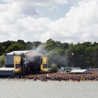 A large crowd of people has gathered by a festival stage on a beach. Some are swimming in the water. In the background there is a forest.