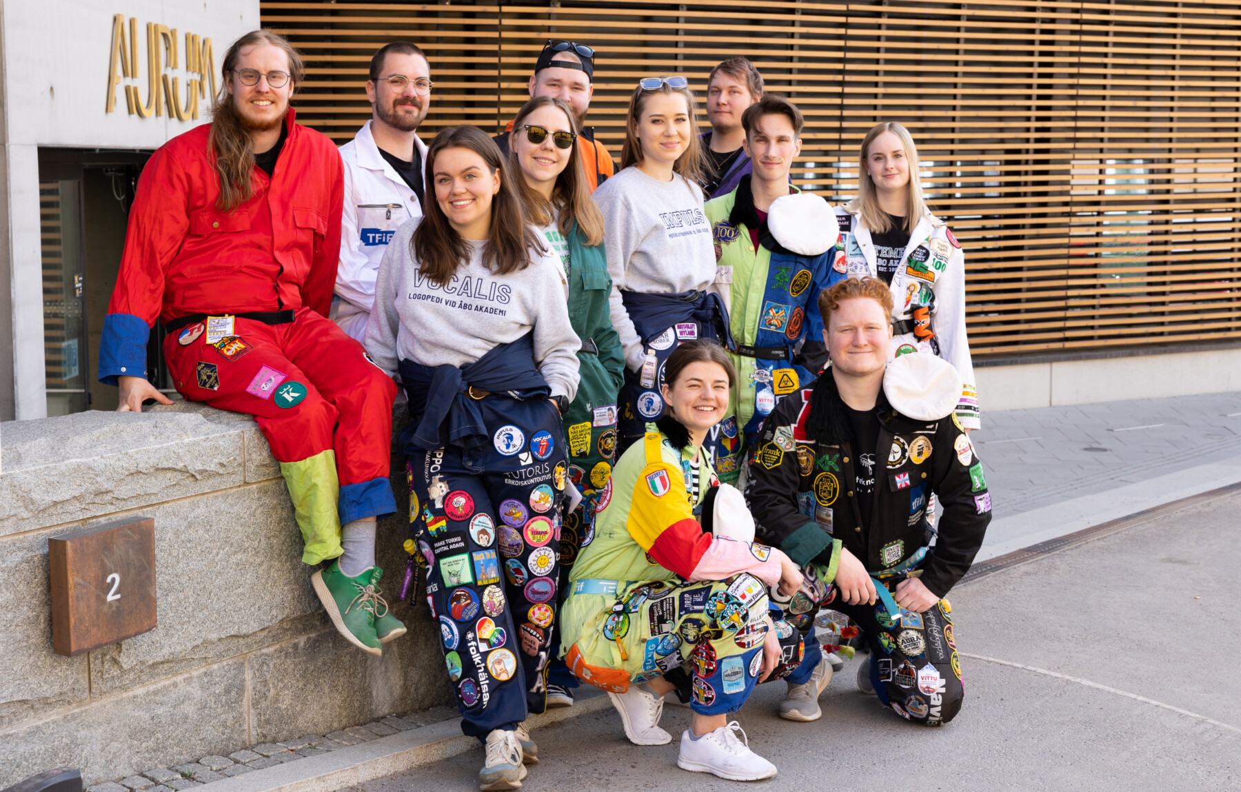 Elf Menschen in bunten Overalls posieren vor einem Universitätsgebäude.