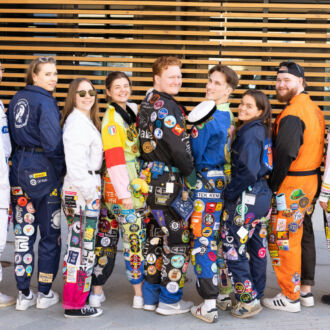 Eleven people wearing decorated overalls stand in a line, looking over their shoulders at the camera.