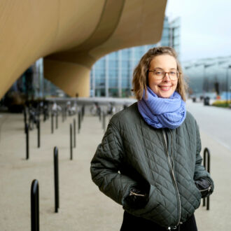 A smiling woman stands beside bike racks in front of a curvy modern wooden building.