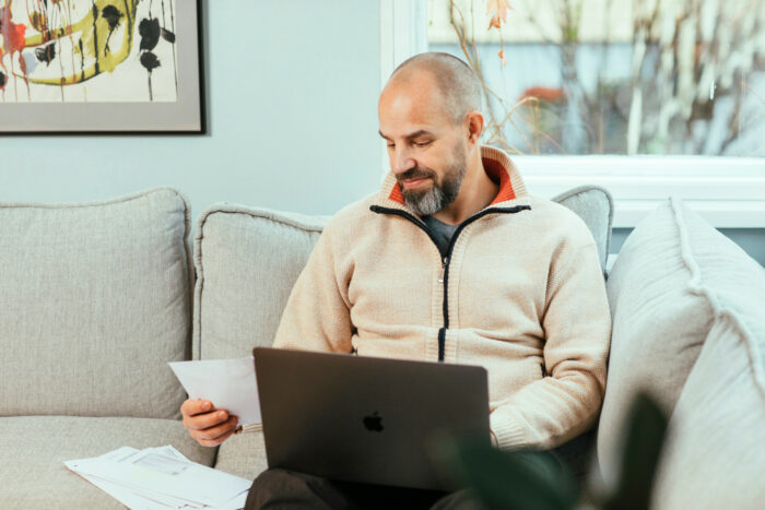 Un homme est installé dans un canapé avec une feuille de papier dans une main et un ordinateur portable posé sur ses genoux.