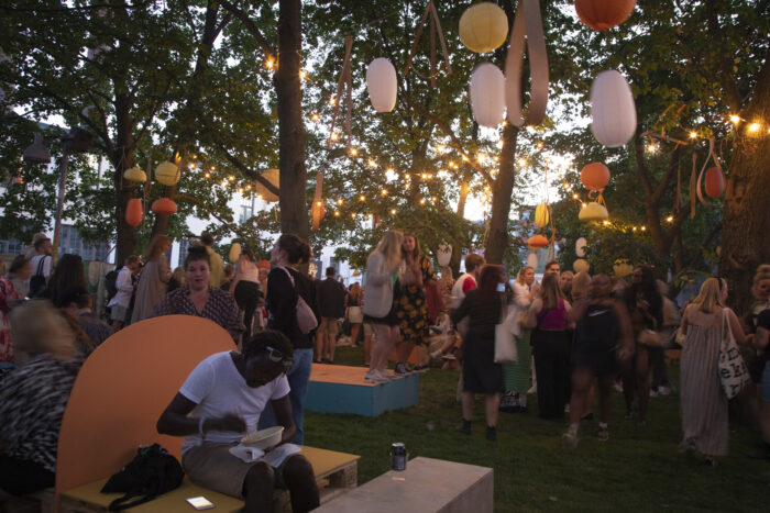 Festivalgoers are talking and dancing in a park-like area with trees and a lawn.