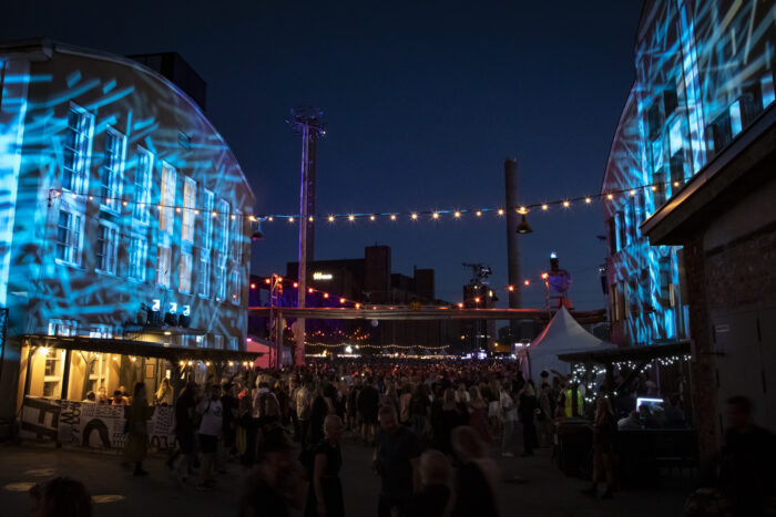 Behind a crowd of concertgoers, buildings are covered in decorative light patterns.