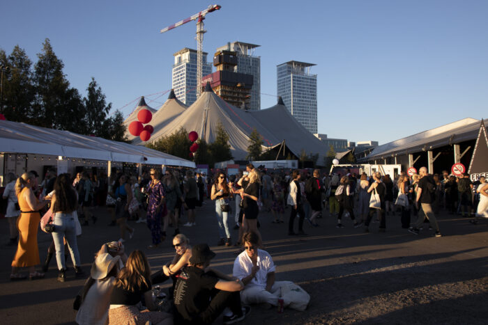 People are milling around on the festival grounds, with three tall buildings visible in the background.