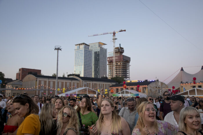Crowds of concertgoers are smiling and talking, with buildings visible in the background.