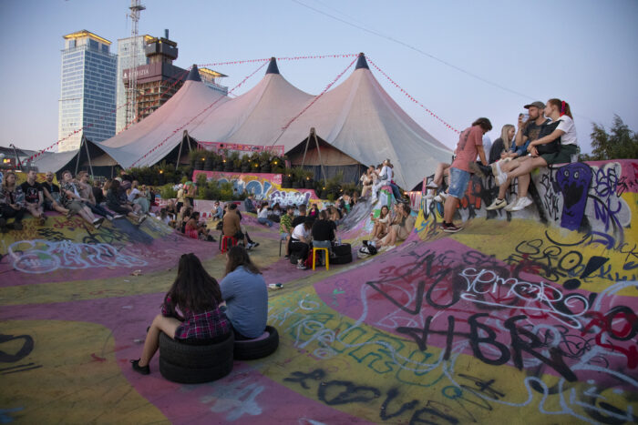 People sit in a colourfully painted asphalt area, with an enormous tent visible in the background.