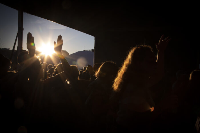 The sun is setting and a pair of hands is sticking up from a concert audience crowd.