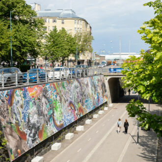 Une peinture murale tout en longueur est visible d'un côté d'une voie bordée d'arbres où marchent deux personnes.