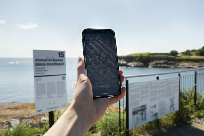 A hand is holding a smartphone up in front of a view that includes islands and the sea.
