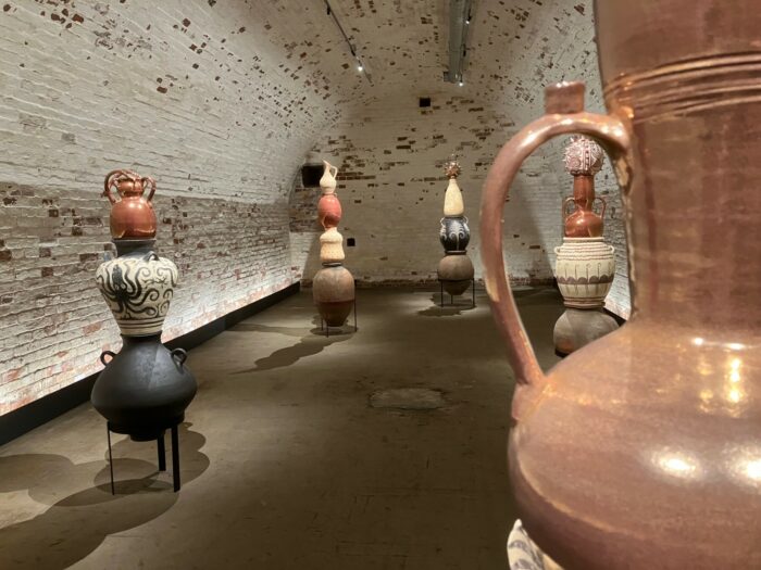 Fantastisch geformte Vasen und Karaffen stehen auf Sockeln in einem Raum.