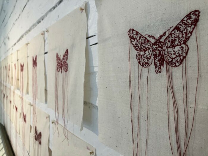 Pedaços de pano estão pendurados na parede, cada um com a imagem de uma mariposa, com fios soltos pendurados na imagem.
