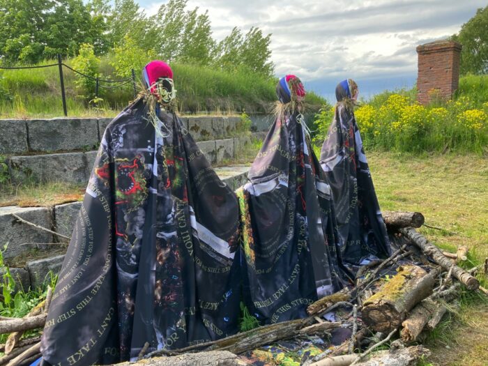 Three human-size figures are draped with dark cloth and appear to have heads made of straw. Trees and flowers are visible in the background.
