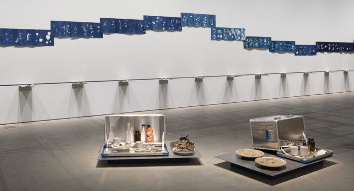 An einer Wand über einer Regalreihe hängen blaue Papierbögen. Davor sind Kisten, Flaschen und Werkzeuge ausgestellt.