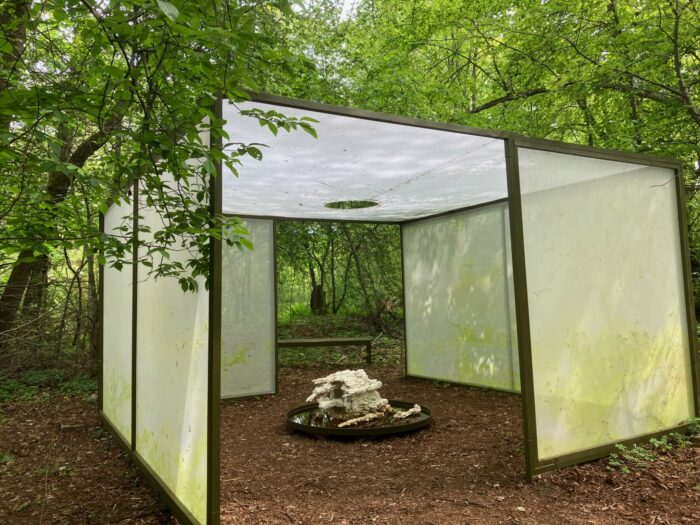 A small abstract plaster sculpture is situated in a forest, partially covered by panels that form a roof and walls.