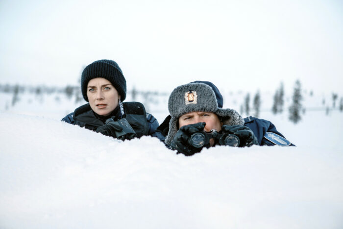Eine Frau und ein Mann mit Wintermützen blicken über den Rand einer Schneewehe.