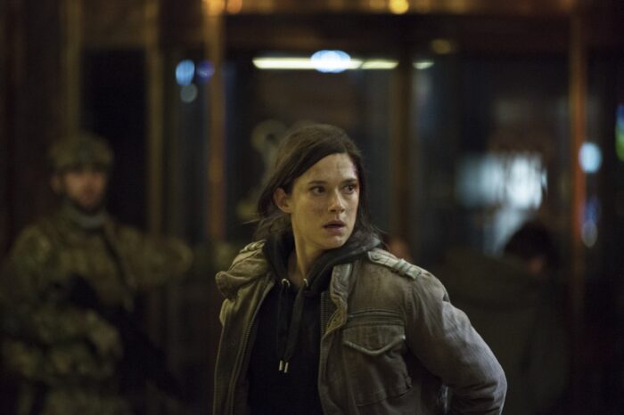 Uma mulher vestindo uma jaqueta militar observa algo fora do enquadramento da foto, com uma expressão estressada no rosto.