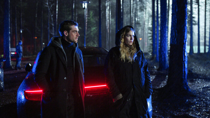 A man and a woman stand by a car in front of a dark forest.
