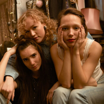 Three teenage girls sit close together, looking into the camera.