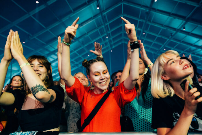 Des festivalières debout au premier rang d’un concert tapent de toutes leurs forces dans leurs mains jointes au-dessus de leurs têtes.