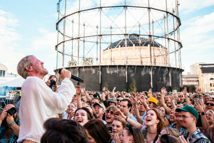 A man in a white shirt sings into a microphone on an outdoor stage while surrounded by cheering fans.