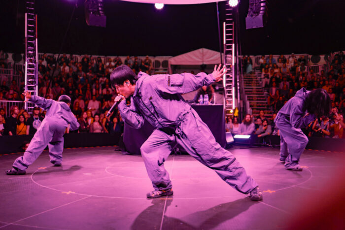 Trois chanteurs habillés de salopettes exécutent des mouvements de danse coordonnés sur une scène baignée de lumière violette.