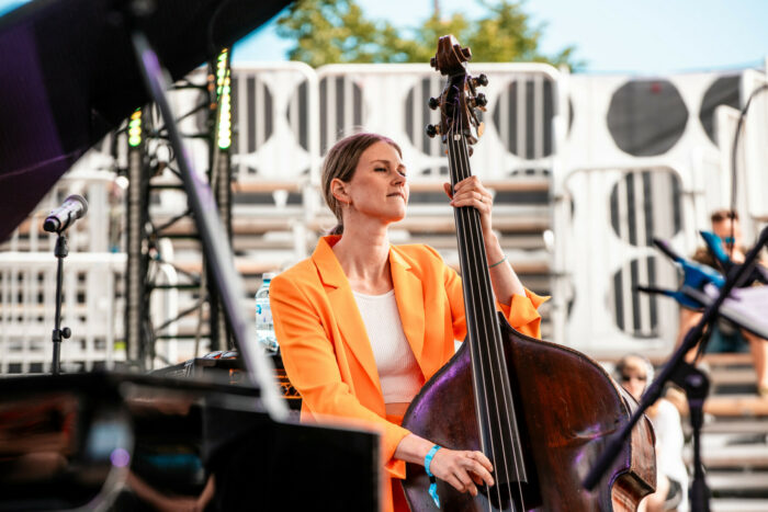 A woman in an orange jacket plays the double bass on an outdoor stage.