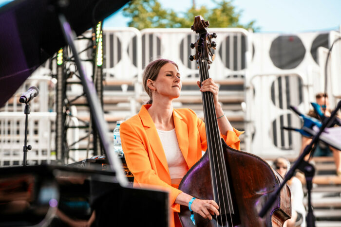 Une femme vêtue d'une veste orange joue de la contrebasse sur une scène en plein air.