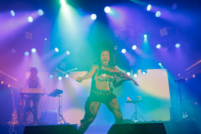 Amid multiple spotlights, a woman holding a violin seems to be shouting something at the audience.