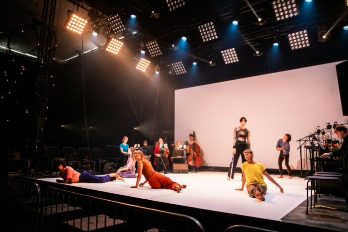 Plusieurs danseurs et danseuses aux tenues vives sont allongés ou se tiennent debout sur une scène flanquée de deux rangées de musiciens.