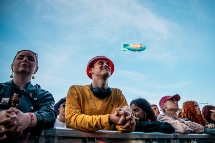 People lean on a railing, while a dirigible is visible far above them in the sky.