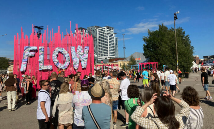 Die Menschen drängen sich um eine hohe rosafarbene Wand, auf der das Wort „Flow“ steht.