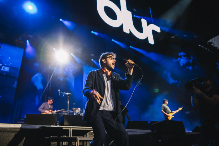 A man sings into a microphone beneath a large sign that says “Blur.”