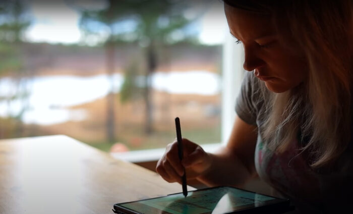 A woman sits at a table, touching an iPad with a stylus, with trees visible outside the window.