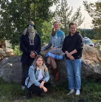 Two women and two men, one of the men wearing a scarf and sunglasses to hide his face, pose in front of a boulder and some trees.