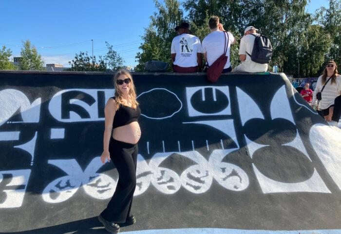 A pregnant woman stands in front of a skateboard ramp where other people are sitting and standing.