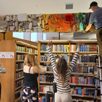 Several students and a man work together on ladders to install a colourful climate art piece made of reused materials above library shelves.