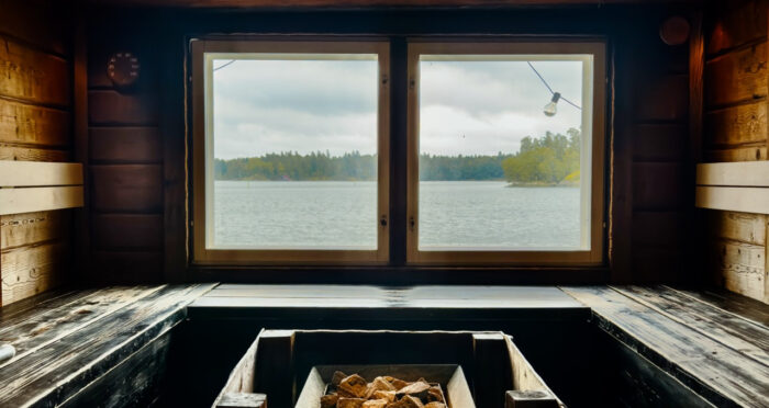 Uma sauna retangular com bancos distribuídos em três paredes, fogão no meio e janelas com vista para o mar.