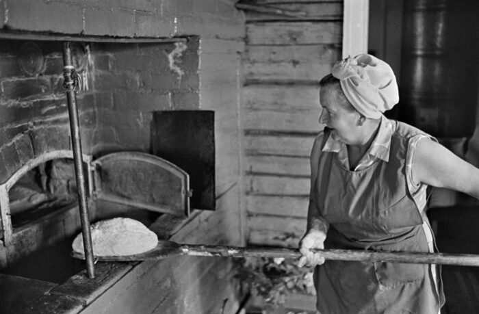 A woman in an apron puts a loaf of bread into a brick oven using a long wooden spatula.