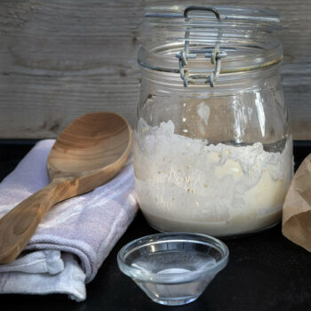 A wooden spoon lies on a tea towel next to a glass jar with several centimetres of a liquid dough mixture in it.